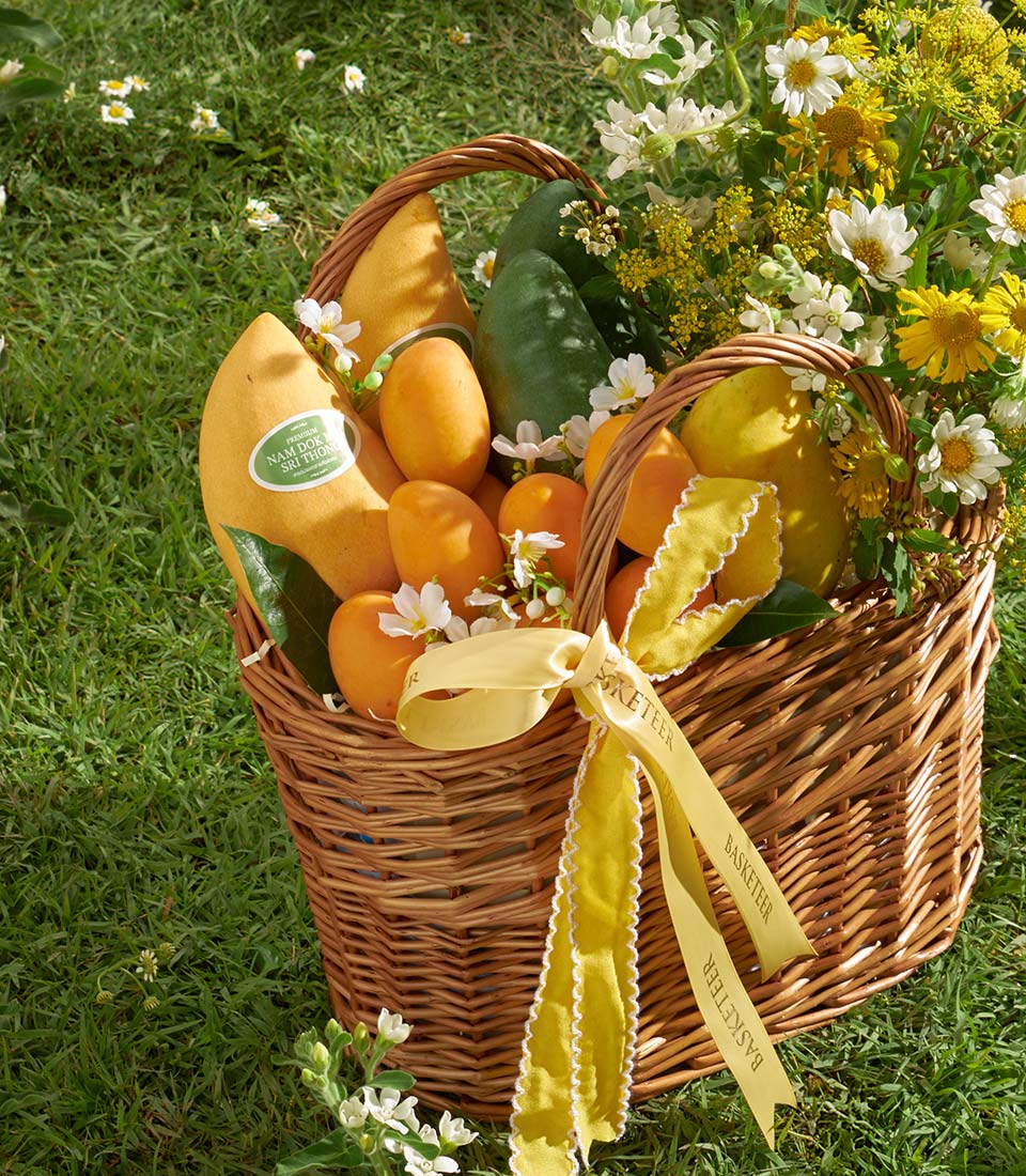 Rustic wicker basket filled with fresh mangoes, tropical fruits, and white wildflowers with yellow ribbon bow