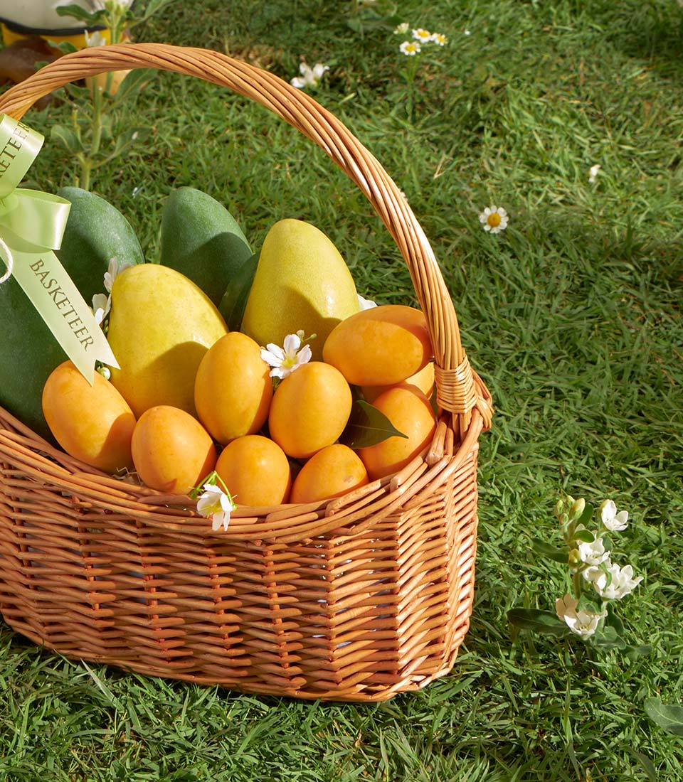 Wicker basket filled with fresh green and golden mangoes, orange marian plums, and white flowers
