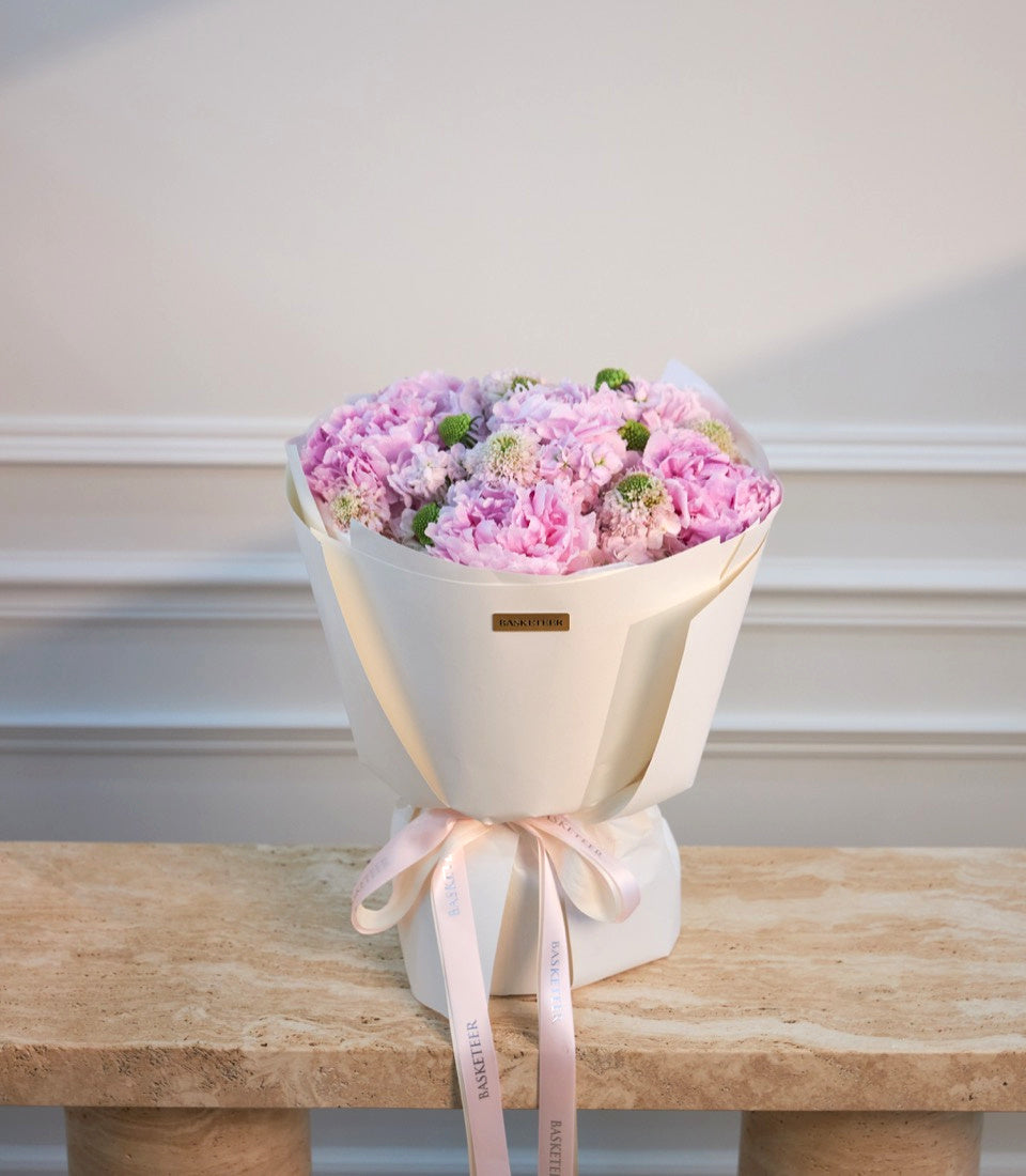 Elegant bouquet of pink and lavender peonies with green hypericum berries wrapped in cream paper with pink ribbon on wooden surface.
