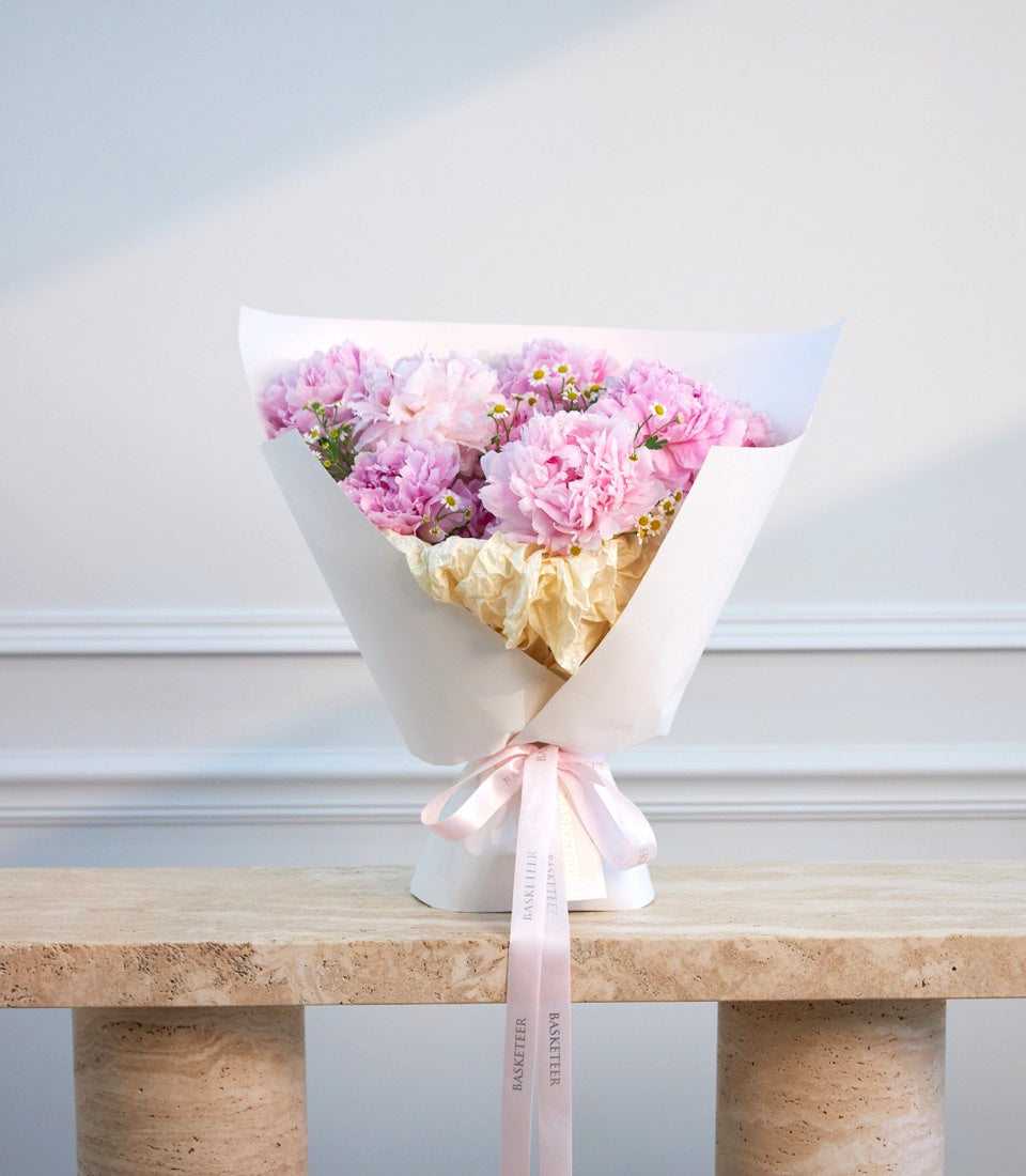 Elegant bouquet of pink peonies and yellow filler flowers wrapped in white paper and gold tissue, tied with a pink ribbon bow.