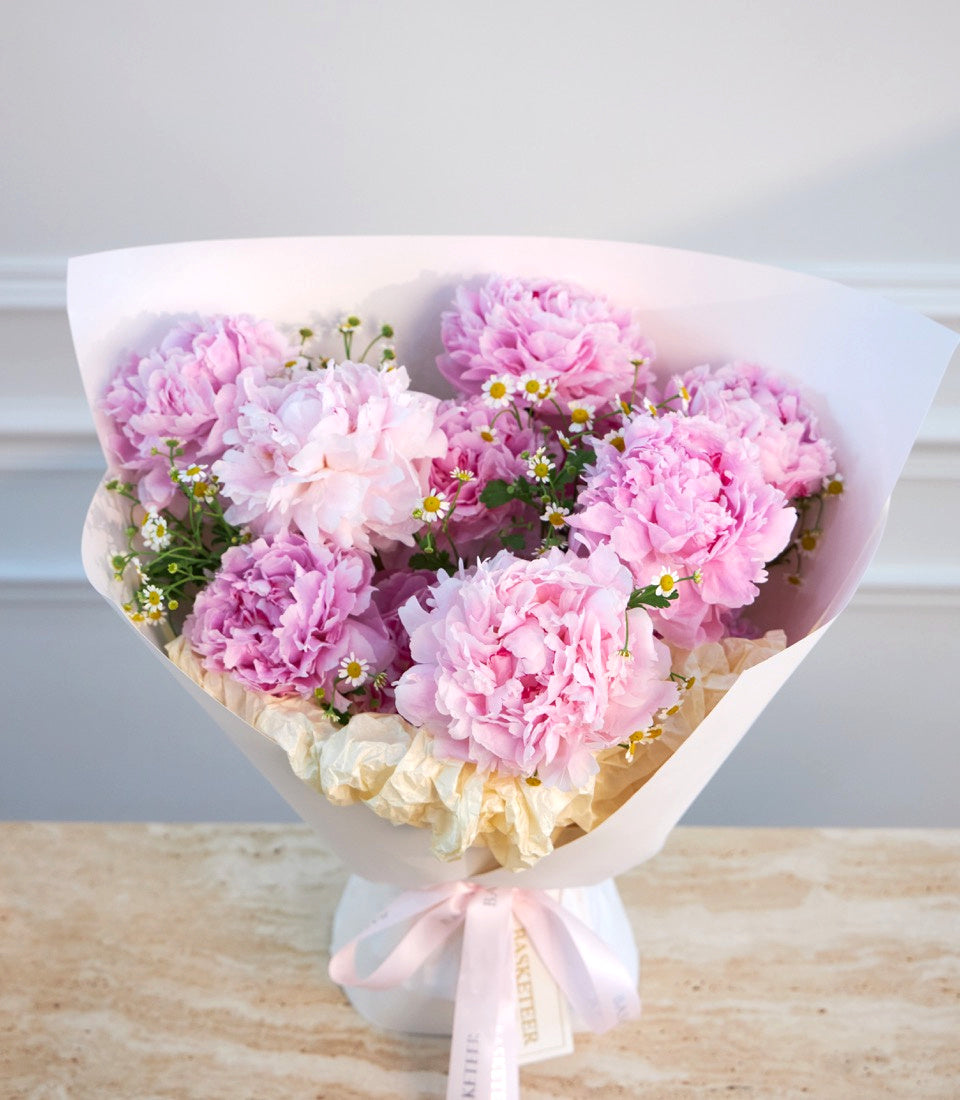 Pink peonies and daisies in white wrapping paper with pink ribbon bow, arranged on wooden surface against white background.