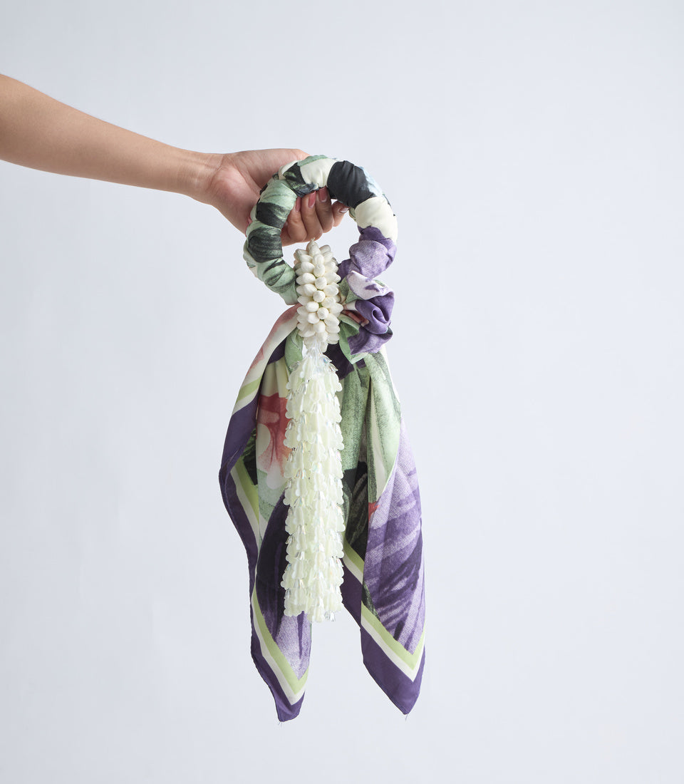 Hand holding a decorative scarf scrunchie with white flower detailing and purple, green, and cream patterned fabric tail.