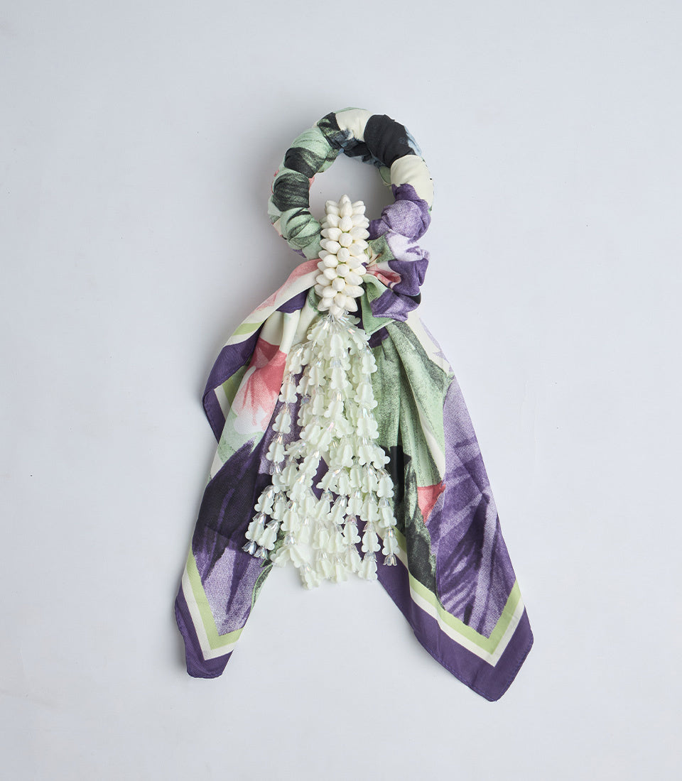 Decorative scarf scrunchie featuring white flower clusters, multicolored plaid fabric tail in purple, green, pink, and cream tones.