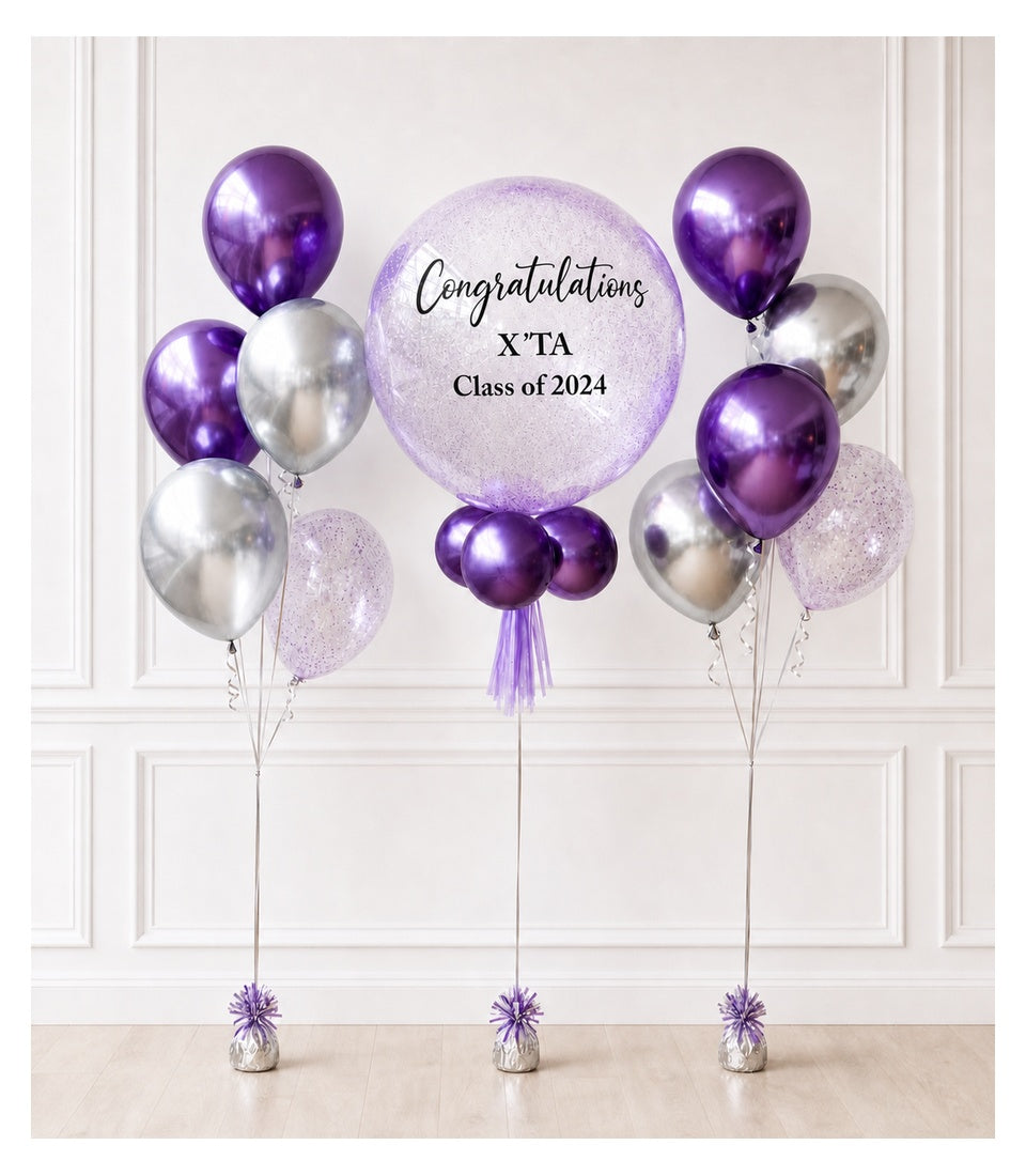Purple and silver metallic balloons with glitter accents arranged around a large purple balloon reading "Congratulations X'TA Class of 2024" against white wall.