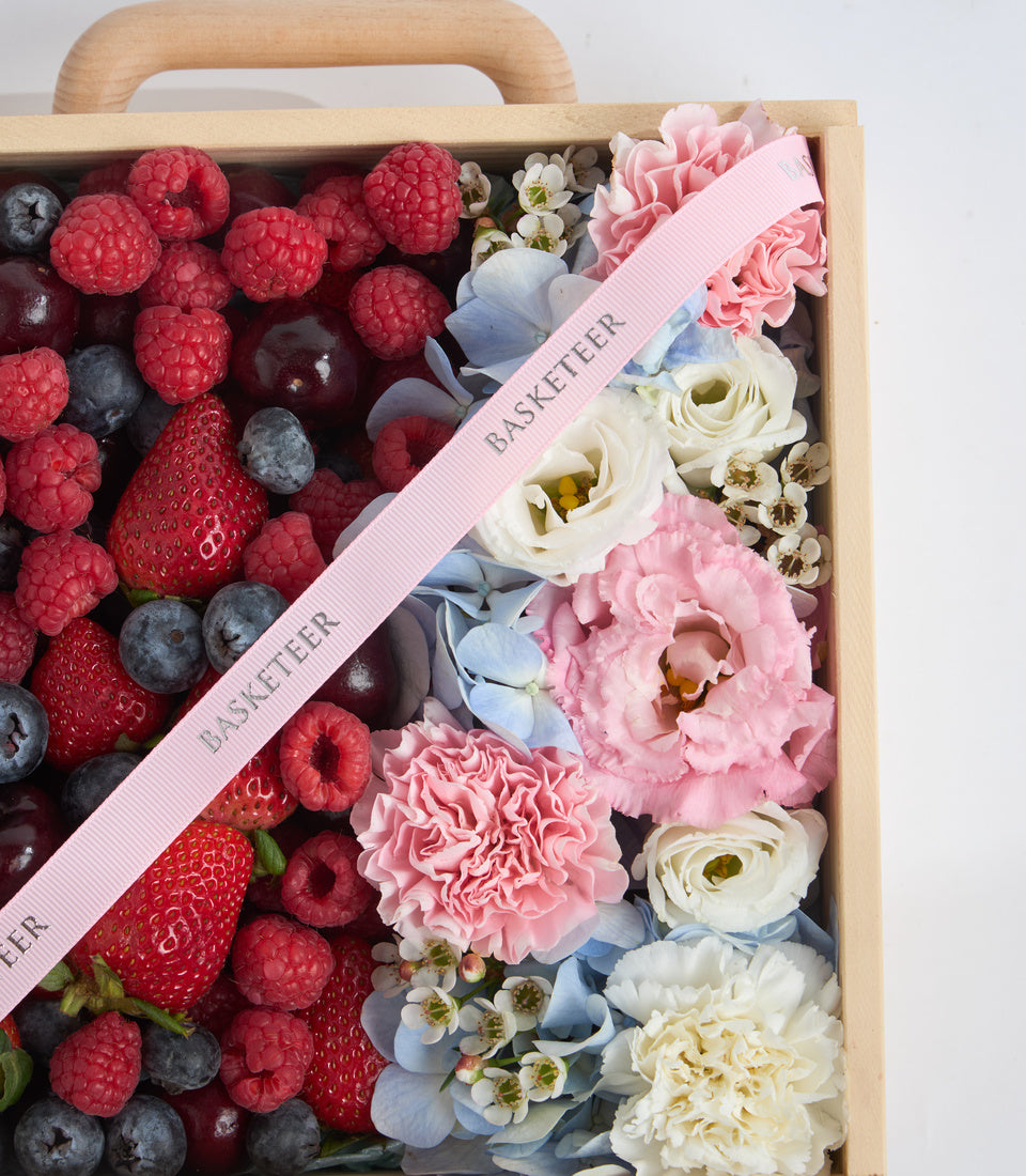 Wooden gift basket with fresh berries, pink and white flowers, blue hydrangeas, and pink "Basketeer" ribbon on white background.