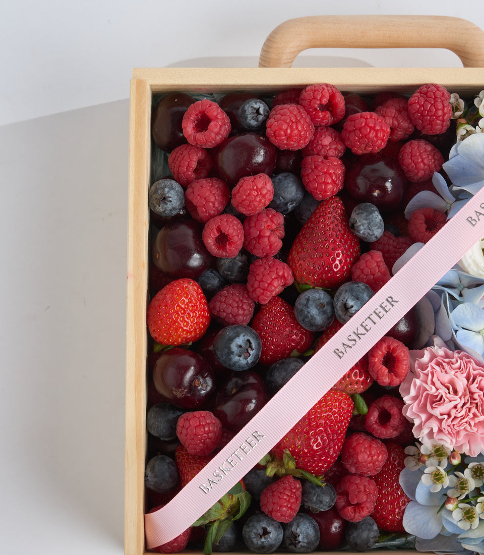 Wooden berry basket filled with fresh raspberries, strawberries, blueberries and cherries, decorated with pink ribbon and flowers.