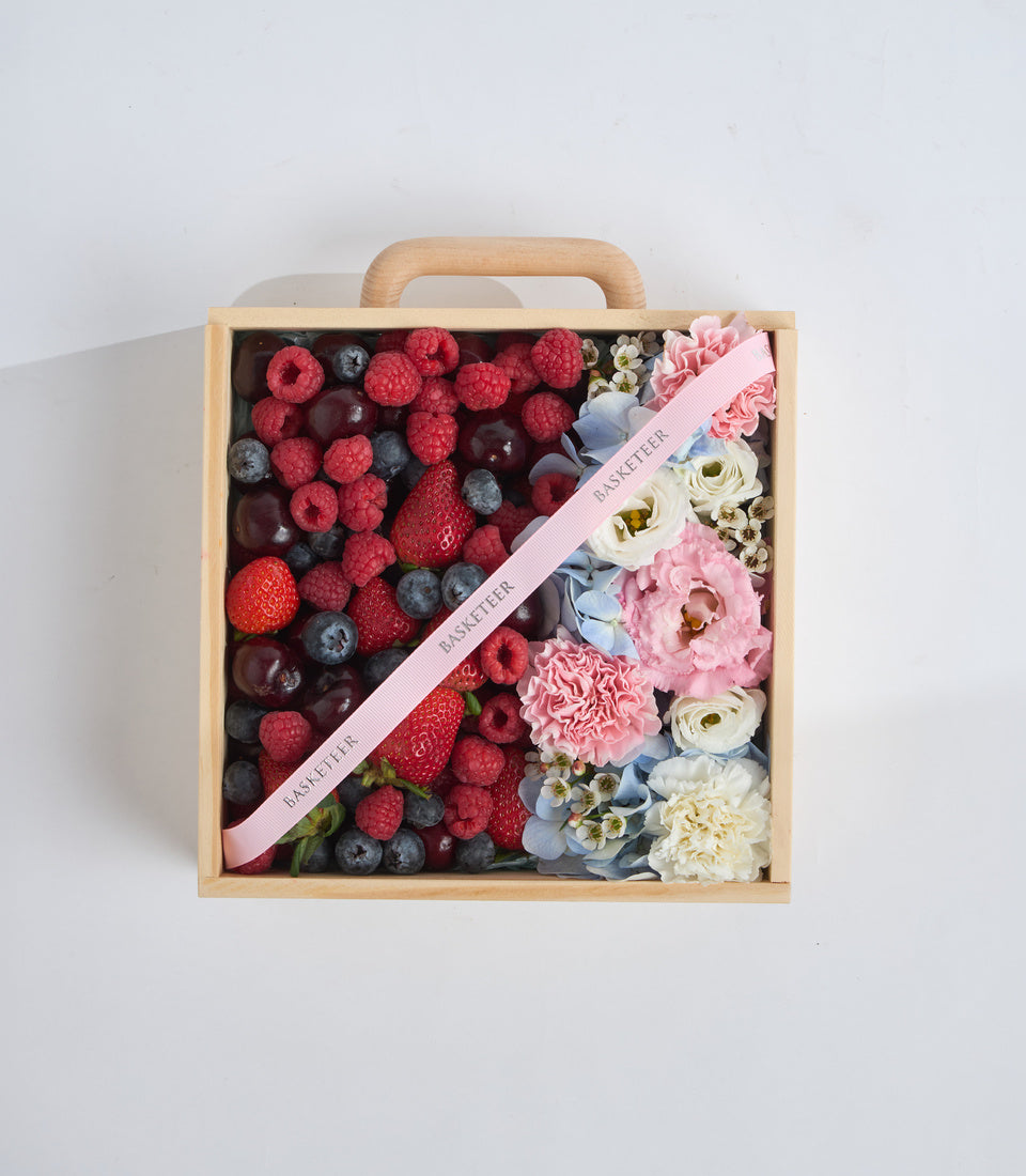 Wooden basket gift box filled with fresh raspberries, strawberries, blueberries, and pink flowers with a pink ribbon on white background.
