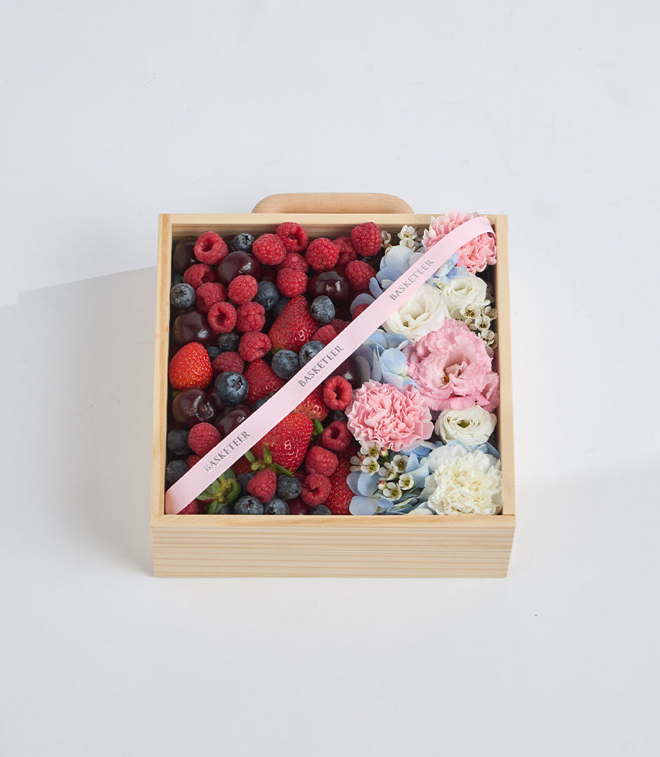 Wooden gift box with fresh berries on left side, pink ribbon, and assorted flowers including pink peonies and white roses on right.