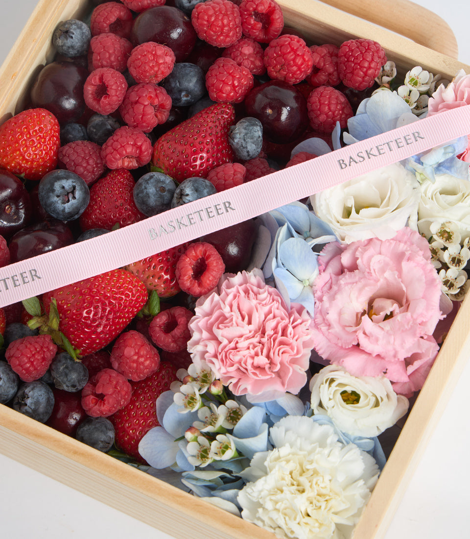 Wooden basket with fresh berries (strawberries, raspberries, blueberries) and pink, white, and blue flowers with "Basketeer" ribbon.