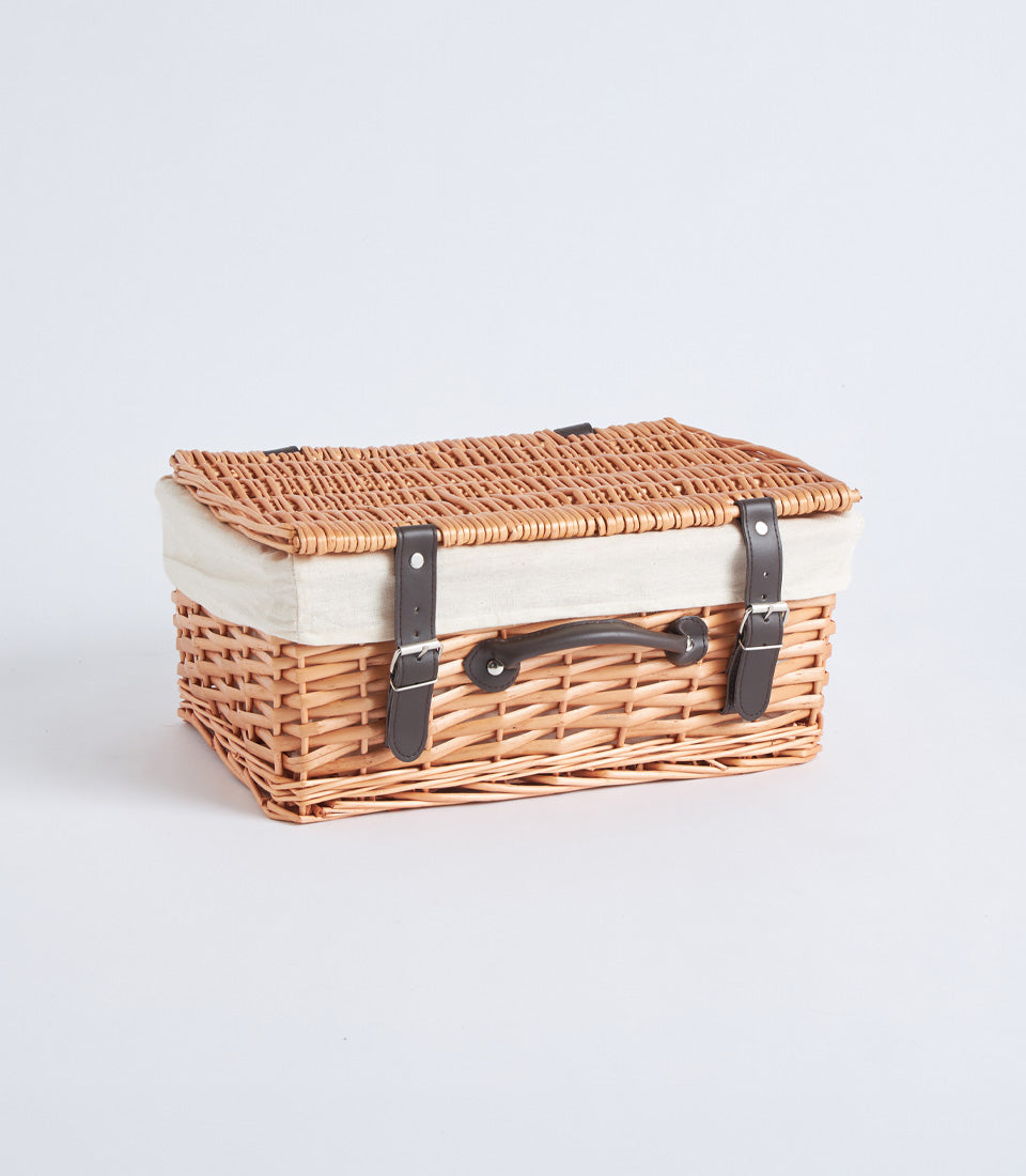 Light brown woven wicker picnic basket with cream linen lining, dark leather straps, and silver buckles