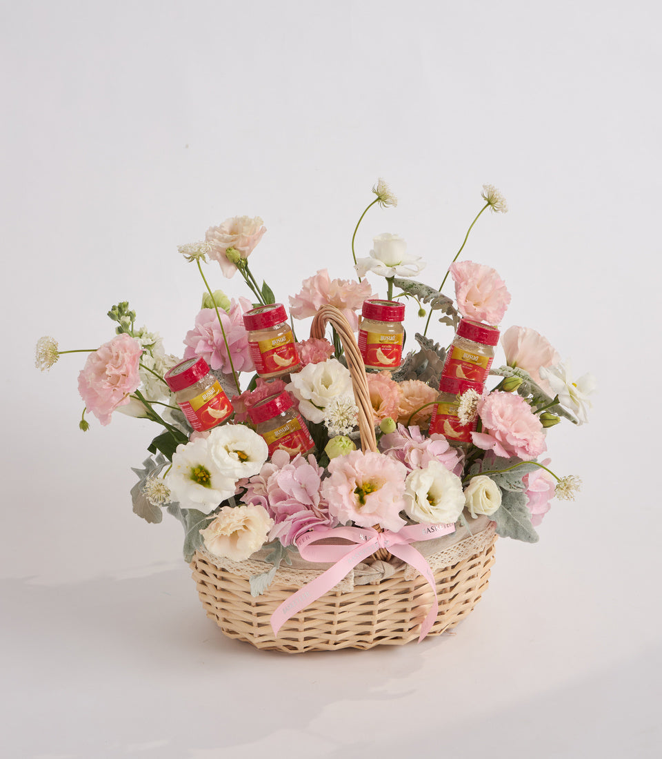 Wicker basket with pink and white carnations, roses, and daisies, accented with red jam jars and a pink ribbon bow.