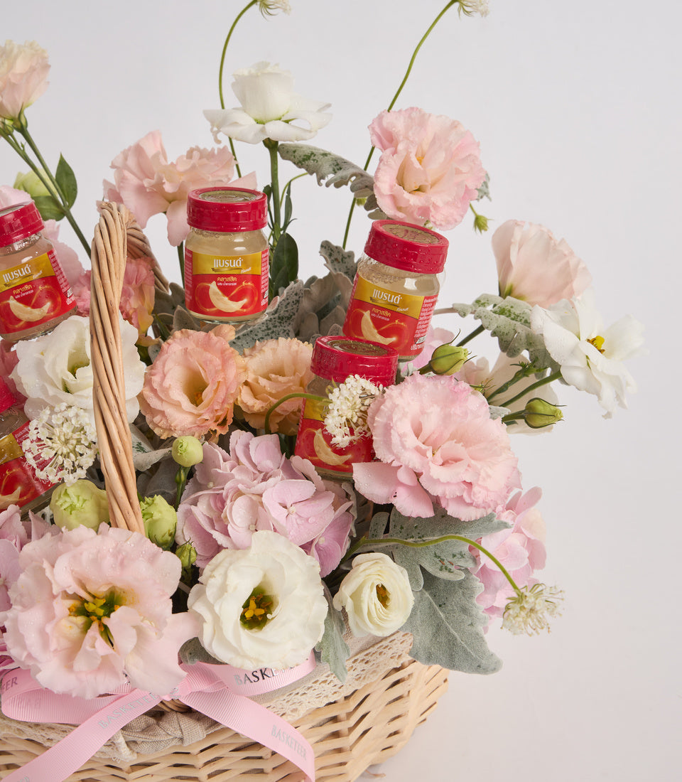 Floral gift basket with pink and white carnations, red product jars, woven basket, grey foliage, and pink ribbon bow on white background.