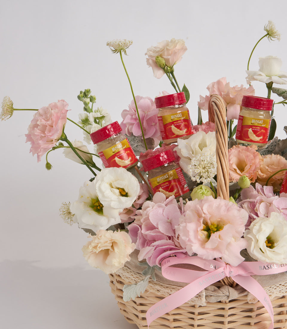 Wicker basket arrangement with pink and white carnations, red product jars with yellow labels, tan rope, and pink ribbon bow on white background.