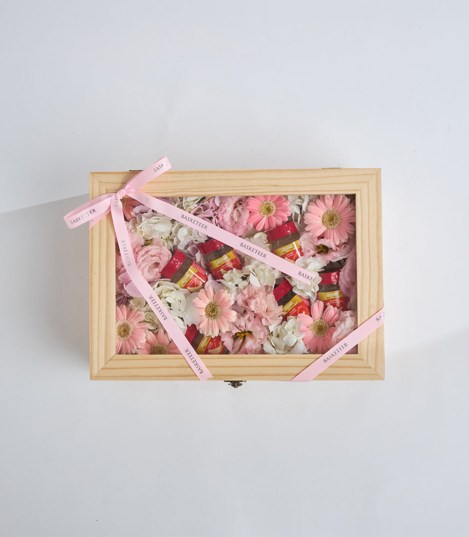 Wooden box filled with pink flowers, white blooms, and red gift bottles, tied with pink "Basketeer" ribbons against white background.
