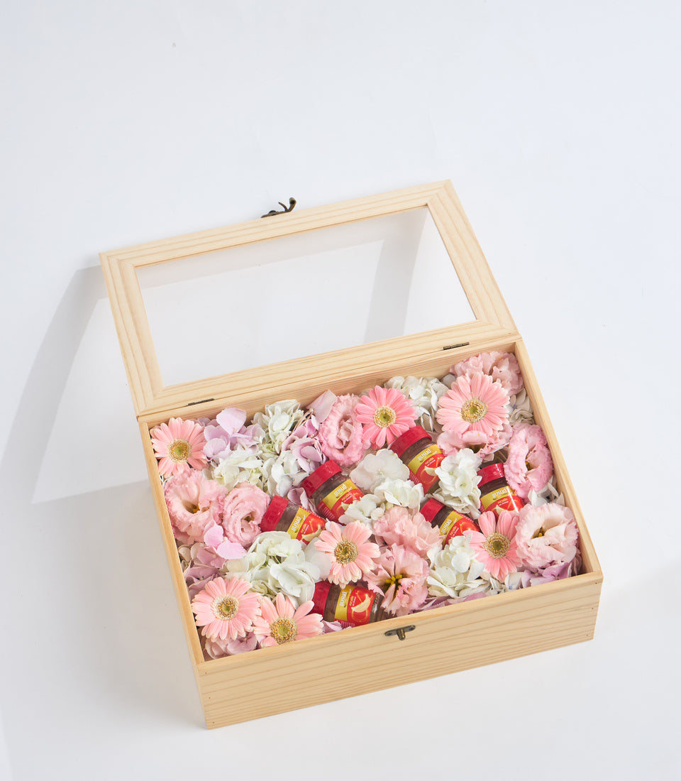 Wooden floral gift box with glass lid containing pink gerberas, white flowers, and red decorative items on white background.