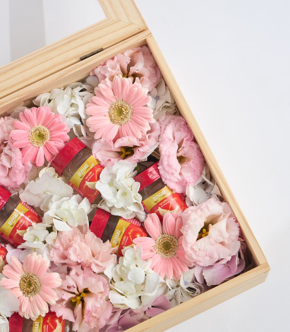 Wooden box filled with pink gerberas, white carnations, pink peonies, and red ribbon-wrapped lip balm tubes arranged as a luxury gift set.