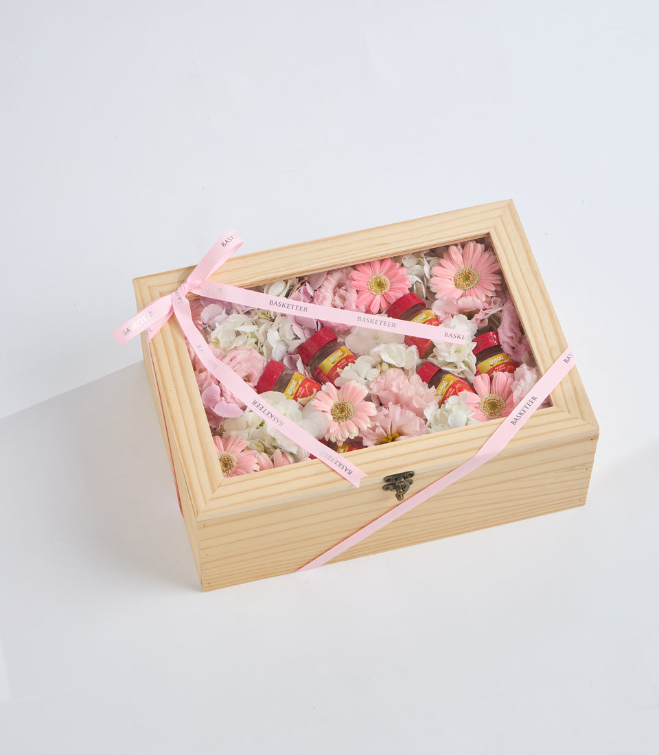 Natural wooden box with glass lid displaying pink gerbera daisies, white flowers, red roses, and gold-trimmed decorative elements, tied with pink ribbon.