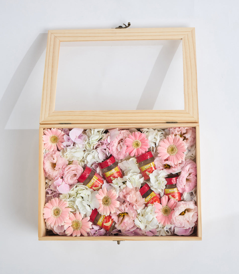 Wooden keepsake box with glass lid displaying pink and white flowers with gold-centered gerberas and small red gift boxes arranged inside.