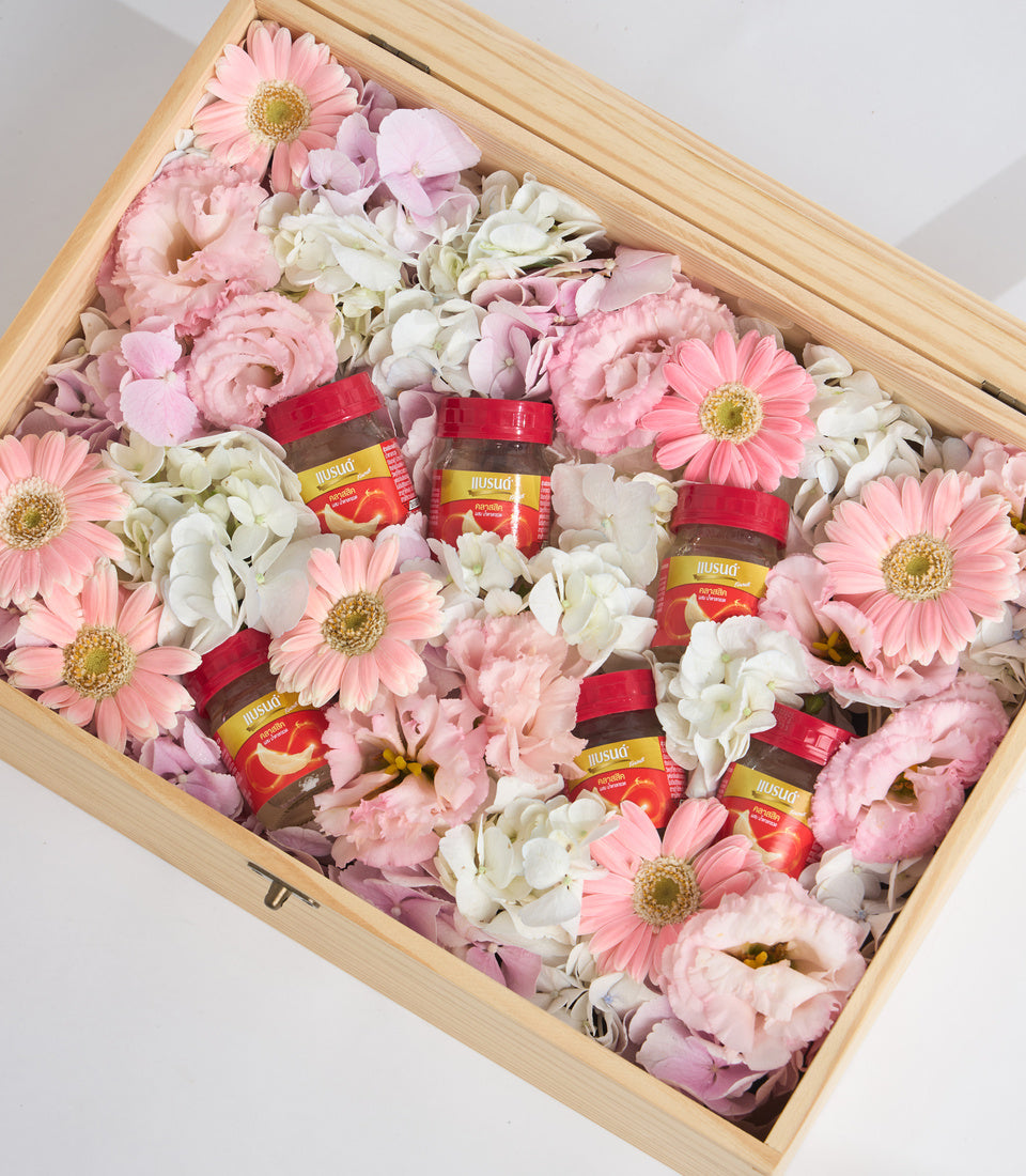 Wooden box filled with pink and white flowers, gerberas, roses, and peonies, nestled with small jars of red-labeled preserves tied with ribbons.
