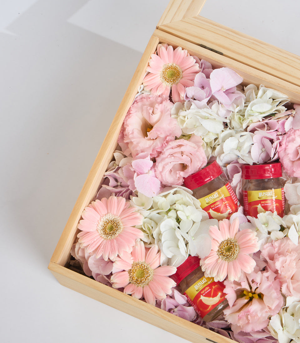 Wooden box filled with pink gerberas, white hydrangeas, carnations, and small jars of red sauce on white background.