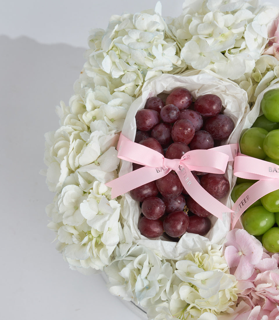 # Alt Text:

Luxe gift arrangement featuring white hydrangeas, pink ribbon-tied pouches of red and green grapes, and blush pink flowers on white background.