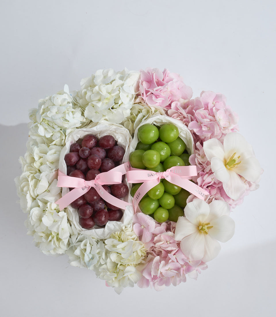 Elegant floral arrangement with white hydrangeas, pink peonies, white anemones surrounding fresh red and green grapes in paper cones with pink ribbons.