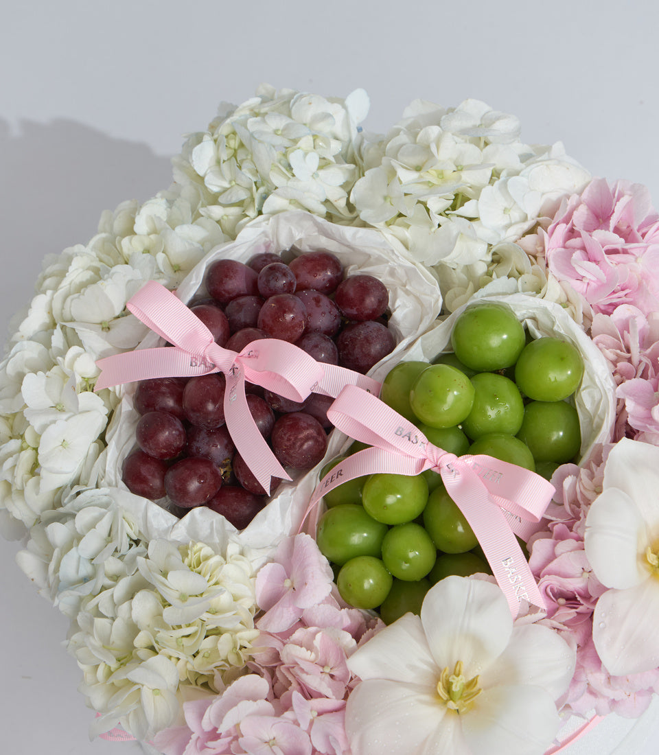Luxury fruit and flower gift arrangement with white and pink peonies, red and green grapes in white boxes with pink ribbons.