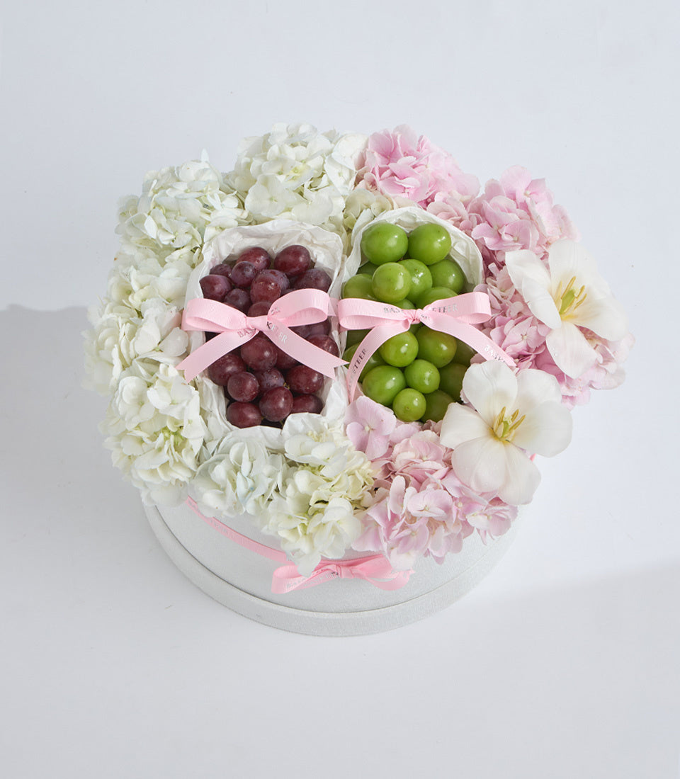 Floral gift arrangement with white and pink hydrangeas, red and green grapes in white pouches with pink ribbons, on white round base.