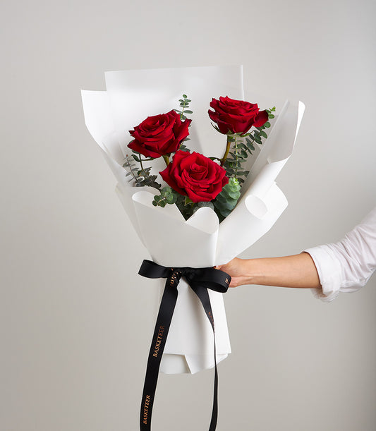 Three vibrant red roses with fresh eucalyptus, elegantly wrapped in white paper and tied with