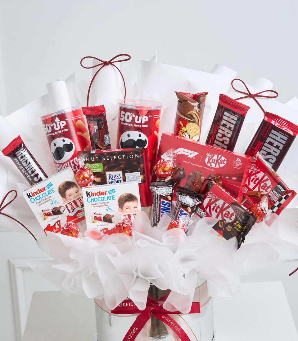 Red and white gift basket filled with chocolates, KitKat bars, Kinder products, and snacks, decorated with white tissue paper and burgundy ribbon bows.