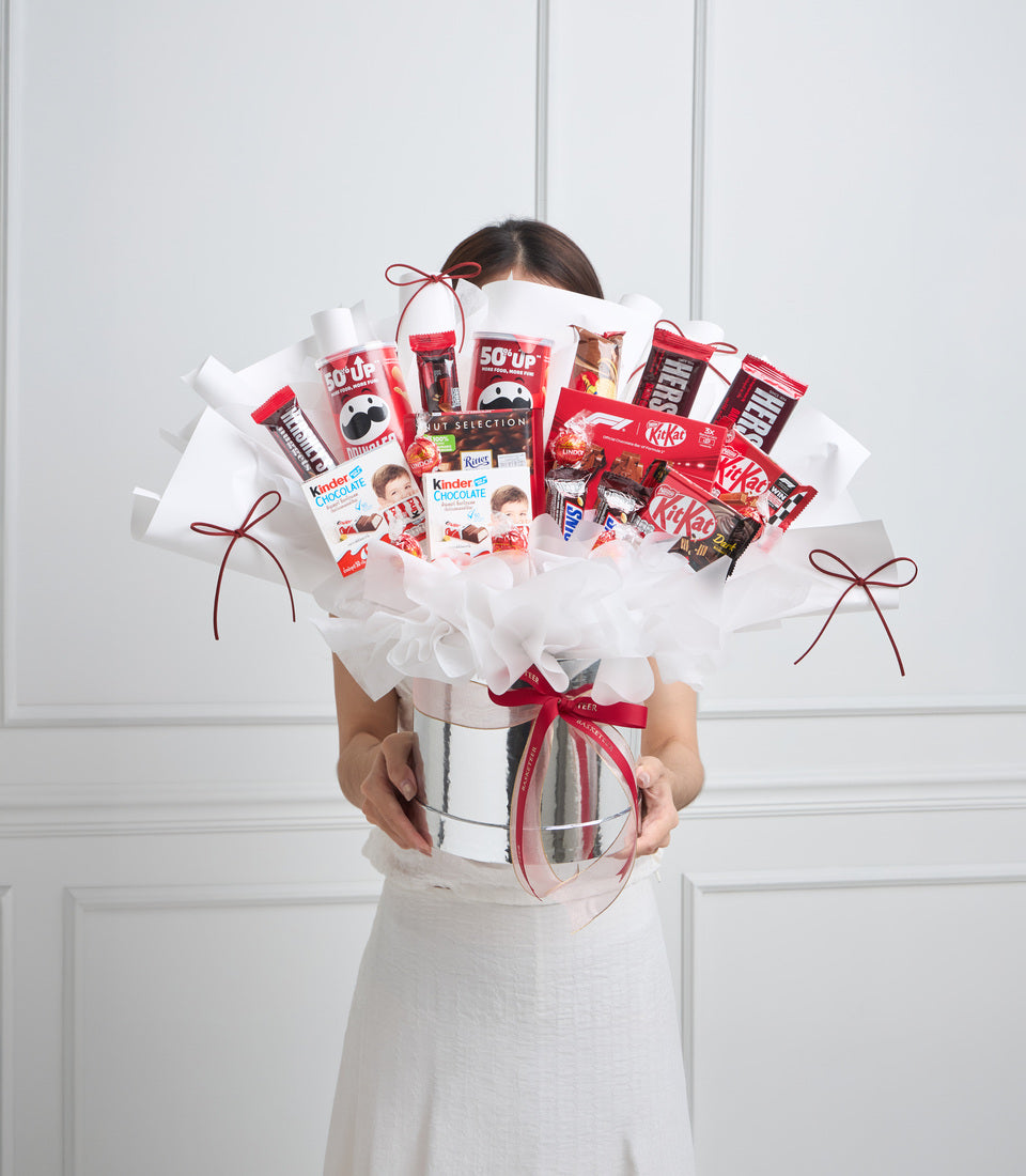 Woman holding large gift bouquet with red and white wrapping, containing chocolate bars, candy, and beverages arranged in cylindrical white container with red ribbon.
