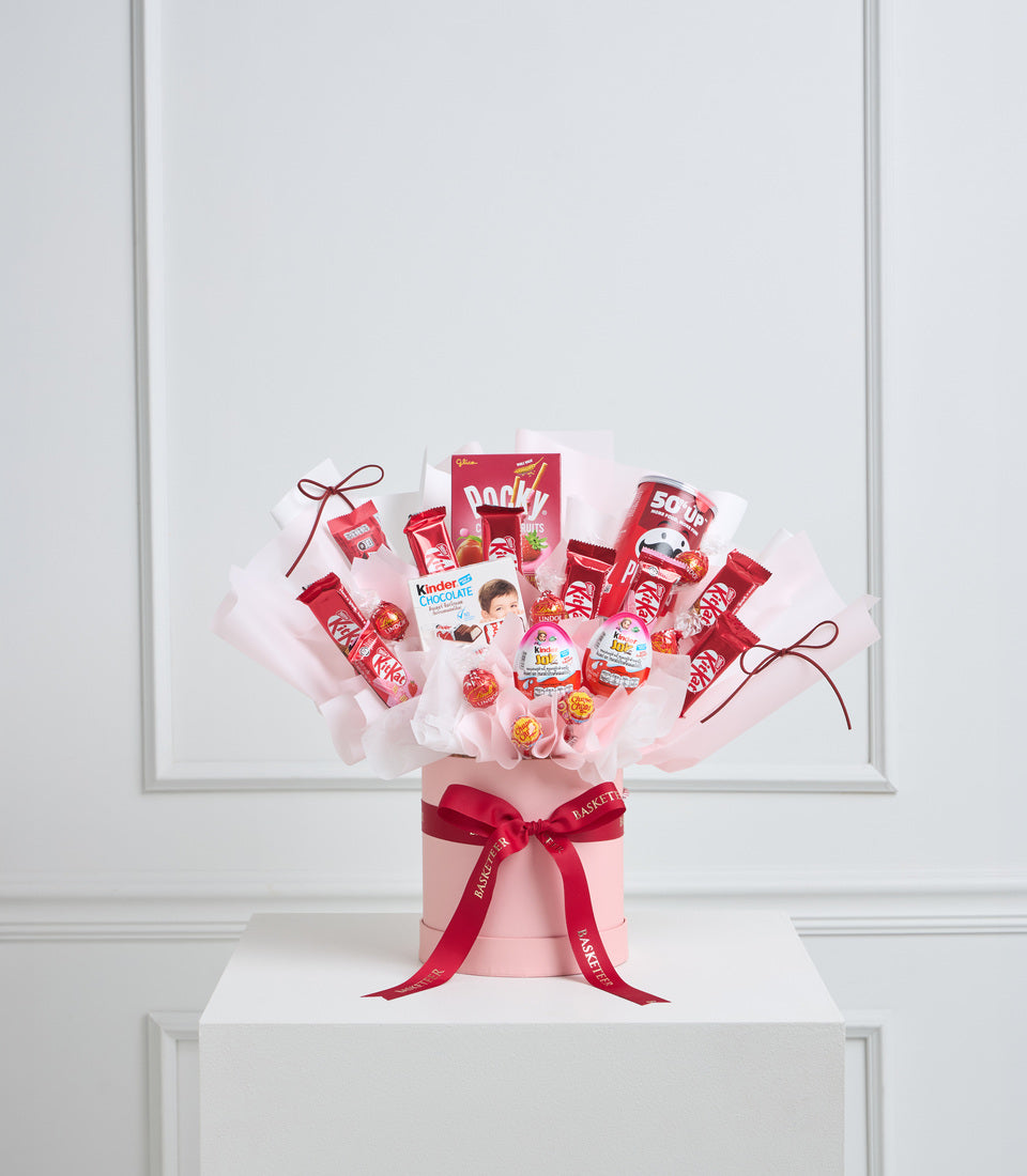 Red and pink candy bouquet arrangement in pink cylindrical box with red ribbon bow, featuring KitKat, Kinder, Pocky and other confectionery items.