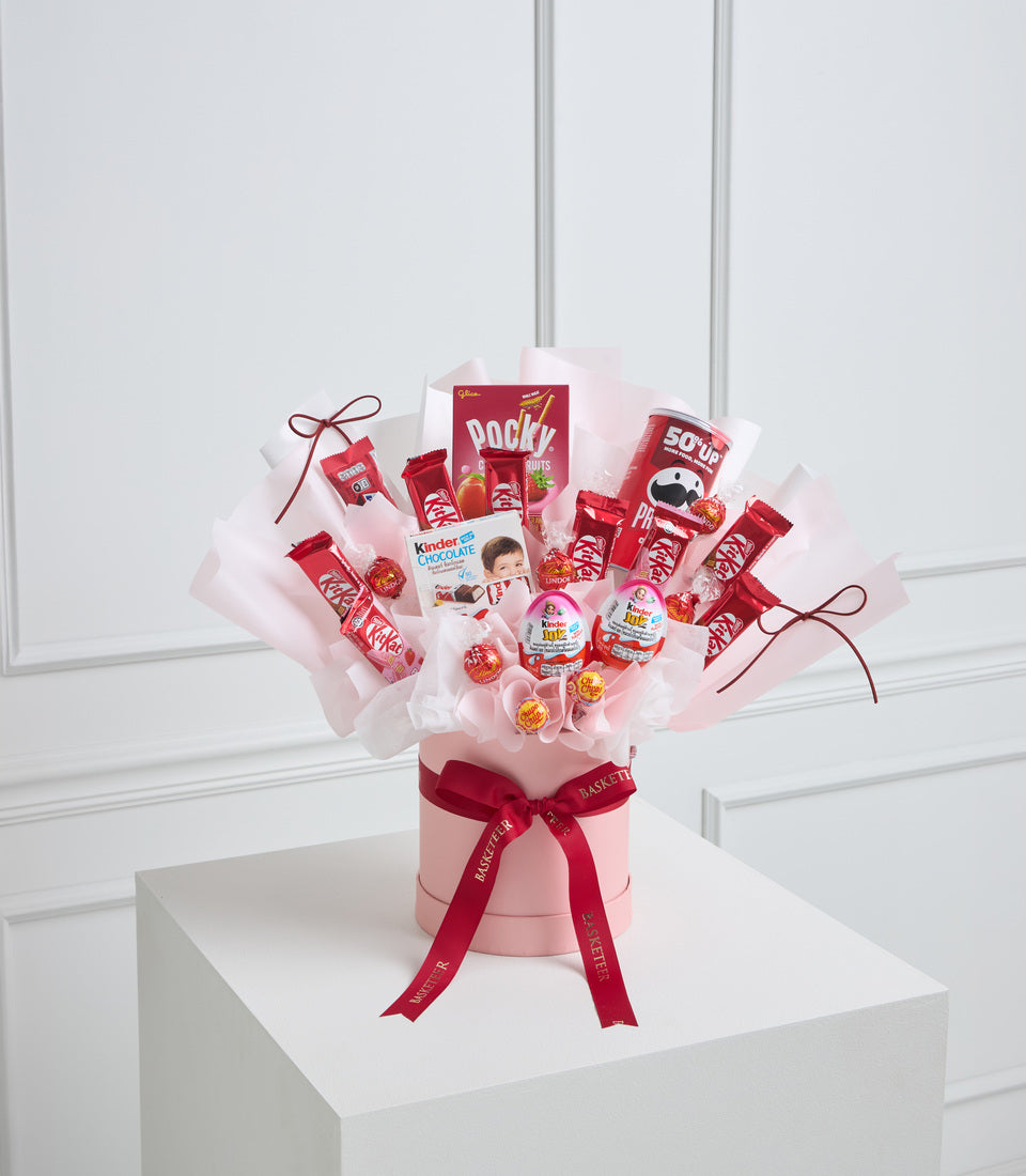 Red and pink candy bouquet arrangement with assorted snacks in pink box with burgundy ribbon bow on white pedestal.