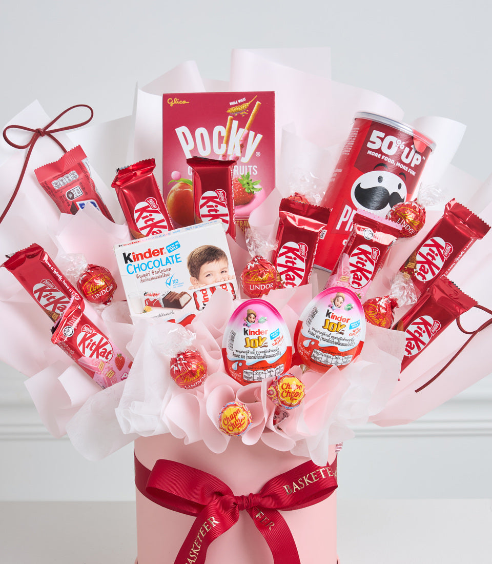 Red and pink candy gift basket with KitKat, Pocky, Kinder chocolates, Chupa Chups lollipops, and Lindor truffles on white tissue paper with red ribbons.