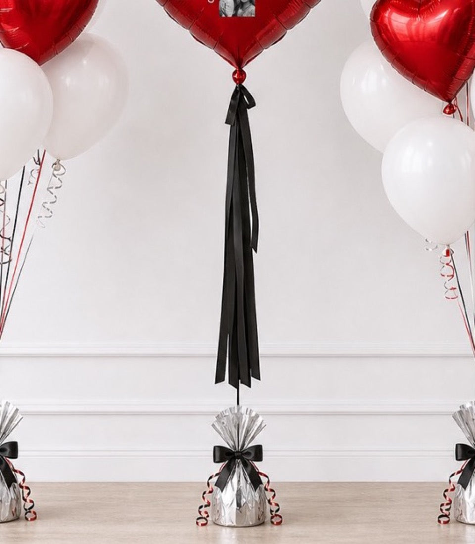 Black tassel balloon weight with silver metallic base, red and white heart-shaped foil balloons, and spiral ribbons in elegant party decoration setup.