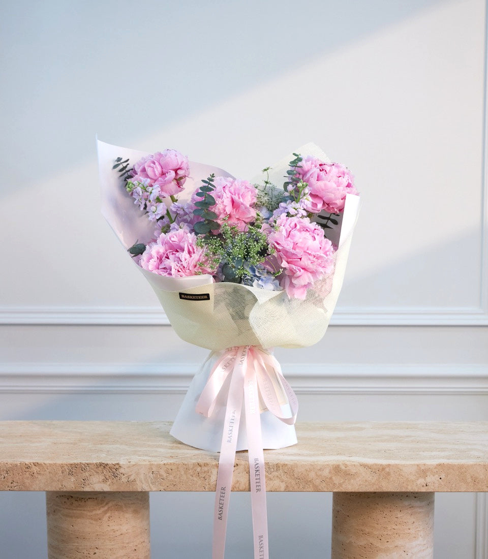 Pink peonies and lavender flowers wrapped in cream textured paper with eucalyptus greenery and pink ribbon bow on wooden surface.