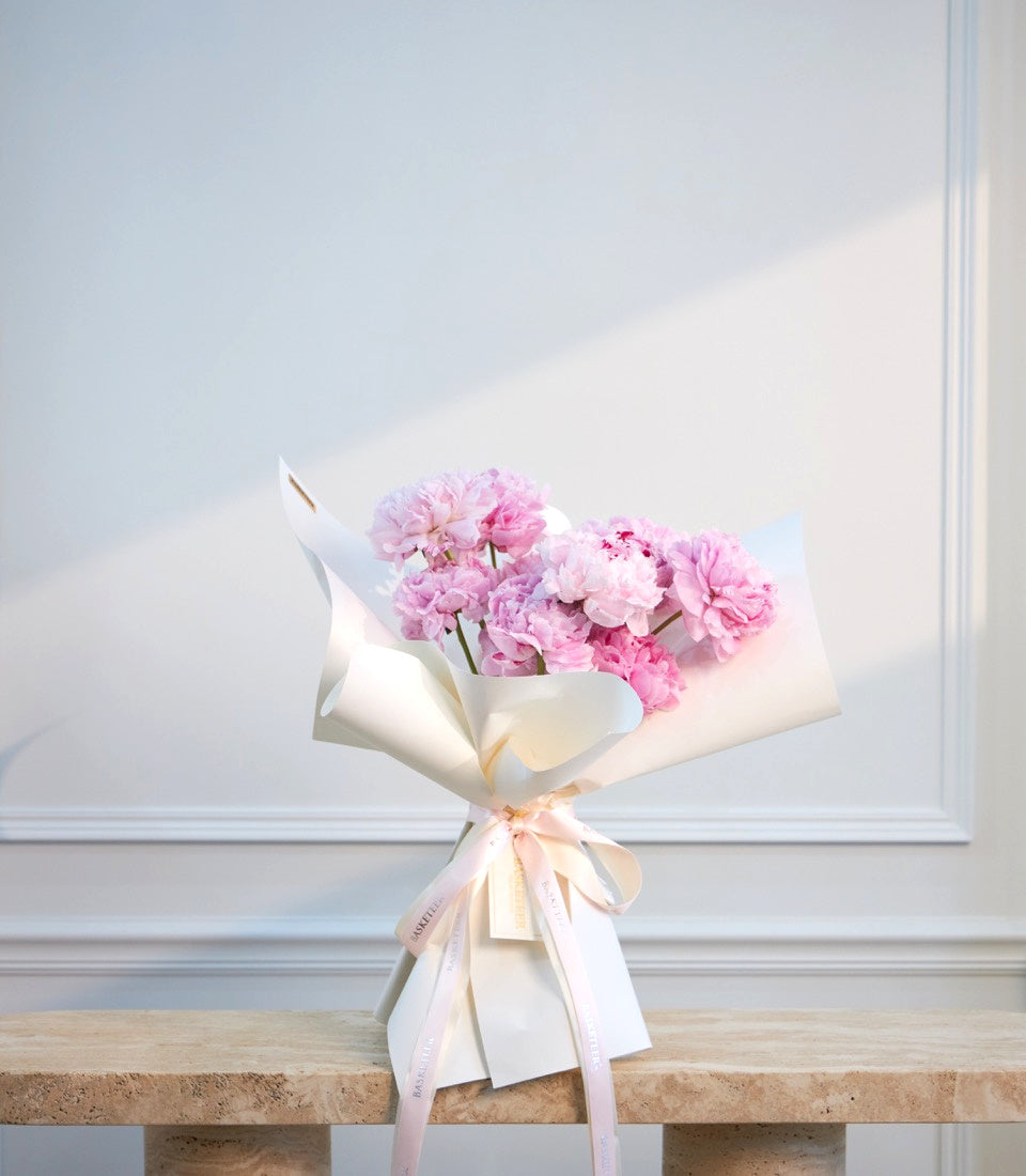 Pink peonies bouquet wrapped in white paper with blush ribbon bow on marble table against white paneled wall.