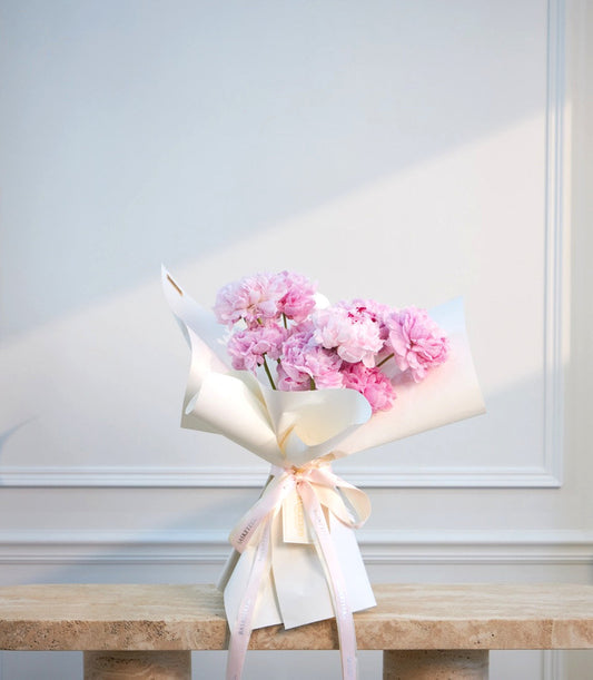 Pink peonies bouquet wrapped in white paper with blush ribbon bow on marble table against white paneled wall.