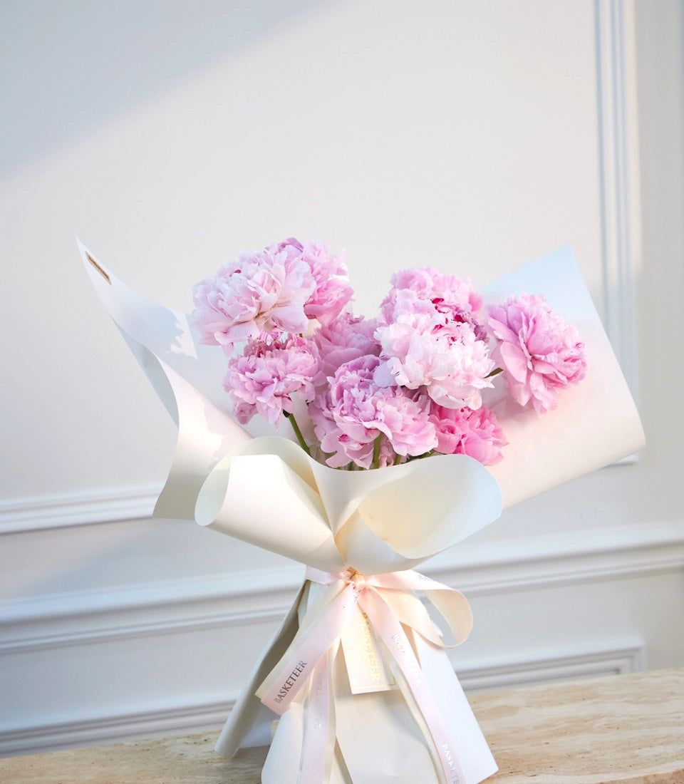 Pink peonies bouquet wrapped in white paper with pale pink ribbon bow, displayed against a bright white interior backdrop.