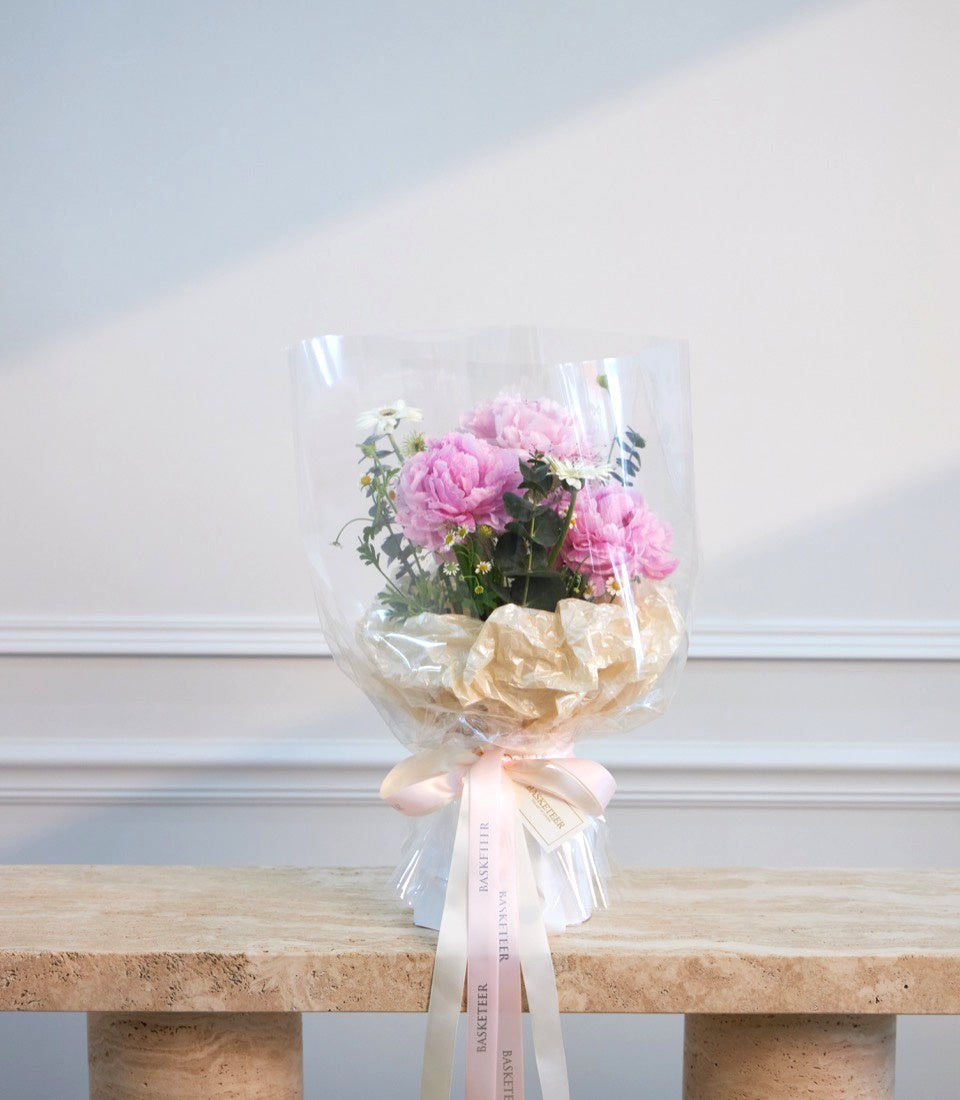 Pink peonies and white flowers wrapped in clear cellophane with cream tissue paper, tied with pink and white ribbons.