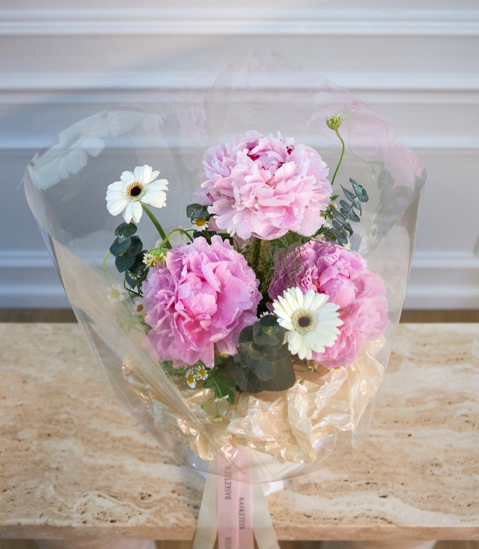 # Alt Text

Wrapped bouquet of pink peonies and white gerbera daisies with greenery, tied with pink ribbon, against a white paneled background.