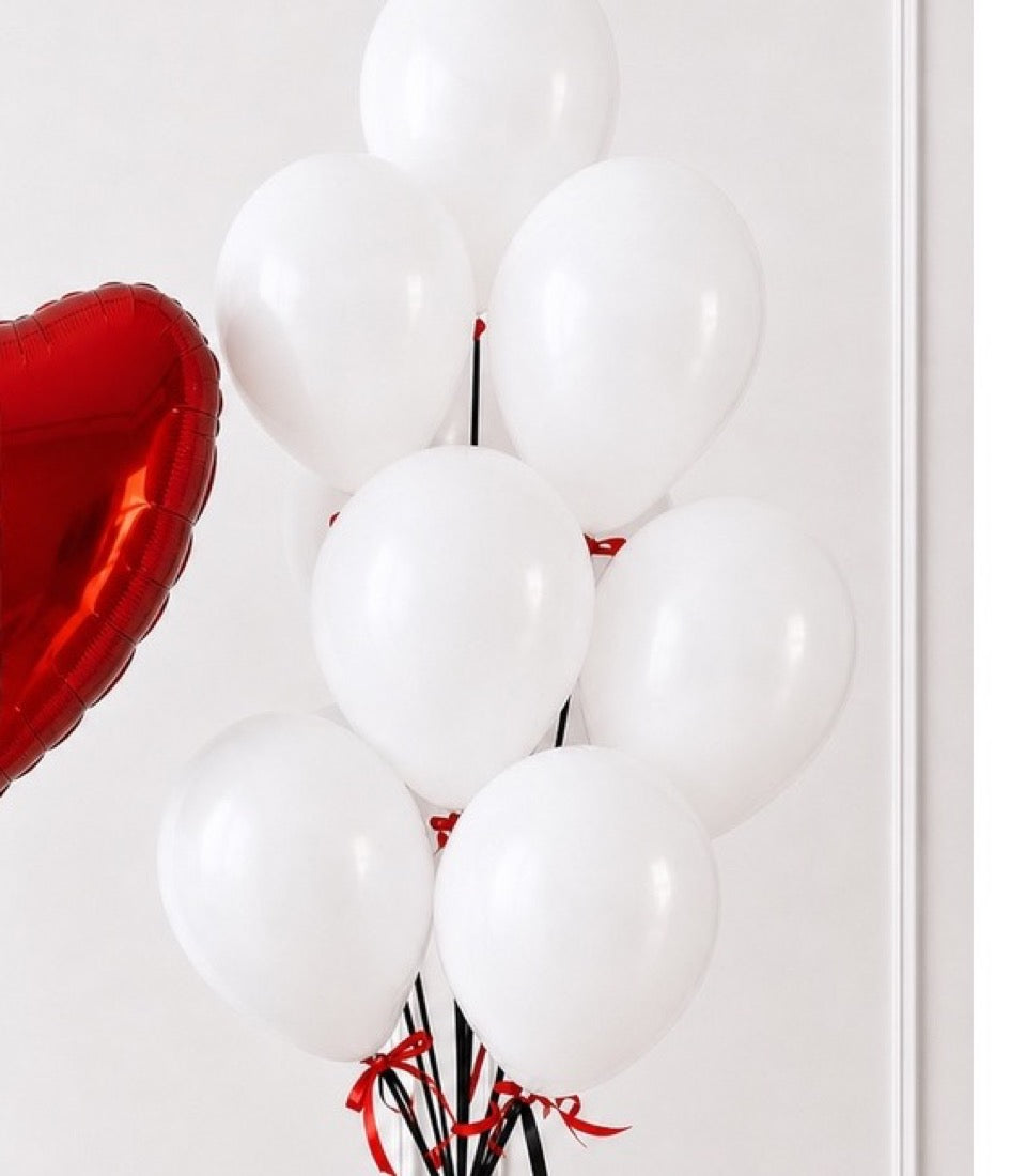 Red heart-shaped foil balloon next to cluster of white round balloons with red and black ribbons against white wall.