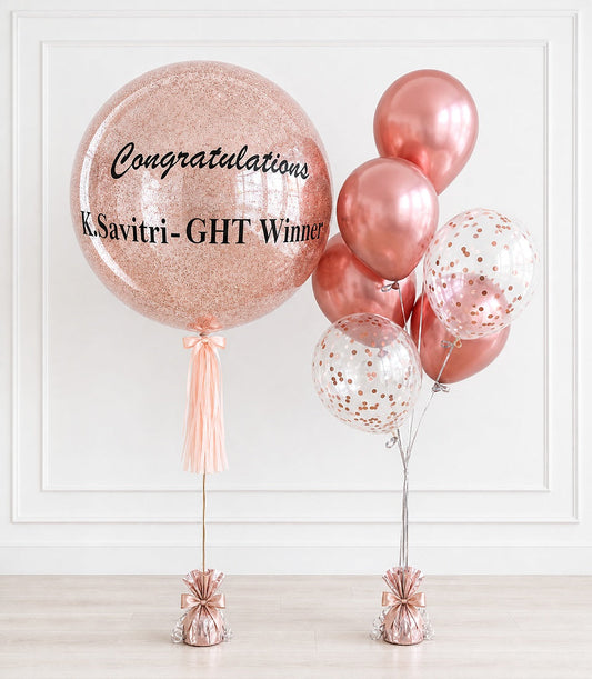 Rose gold glitter balloon with "Congratulations k.Savitri-GHT Winner" text, paired with metallic pink and confetti balloons with tassel weights.