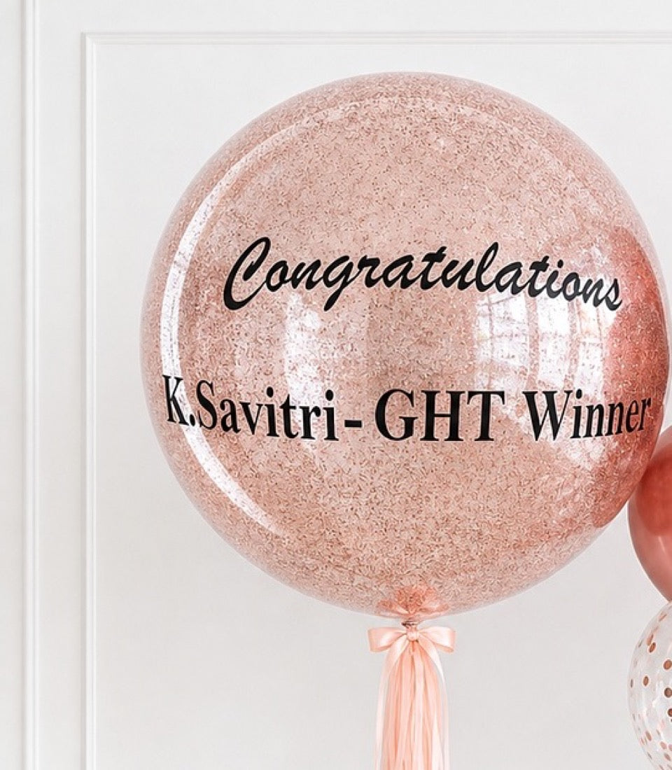 Rose gold glitter balloon with "Congratulations K.Savitri-GHT Winner" text and peach ribbon tassel.