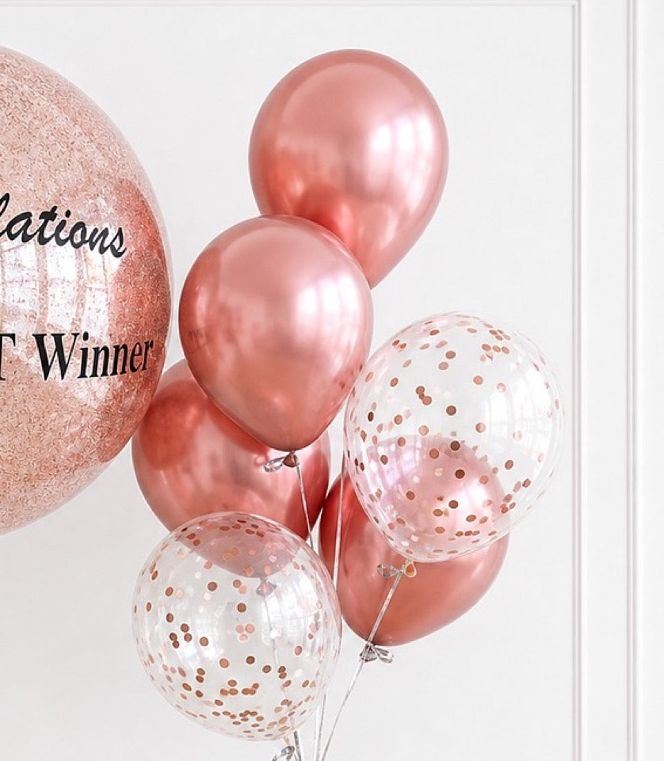 Rose gold balloon cluster featuring metallic and confetti-filled balloons with a glittery "Winner" text balloon against white background.