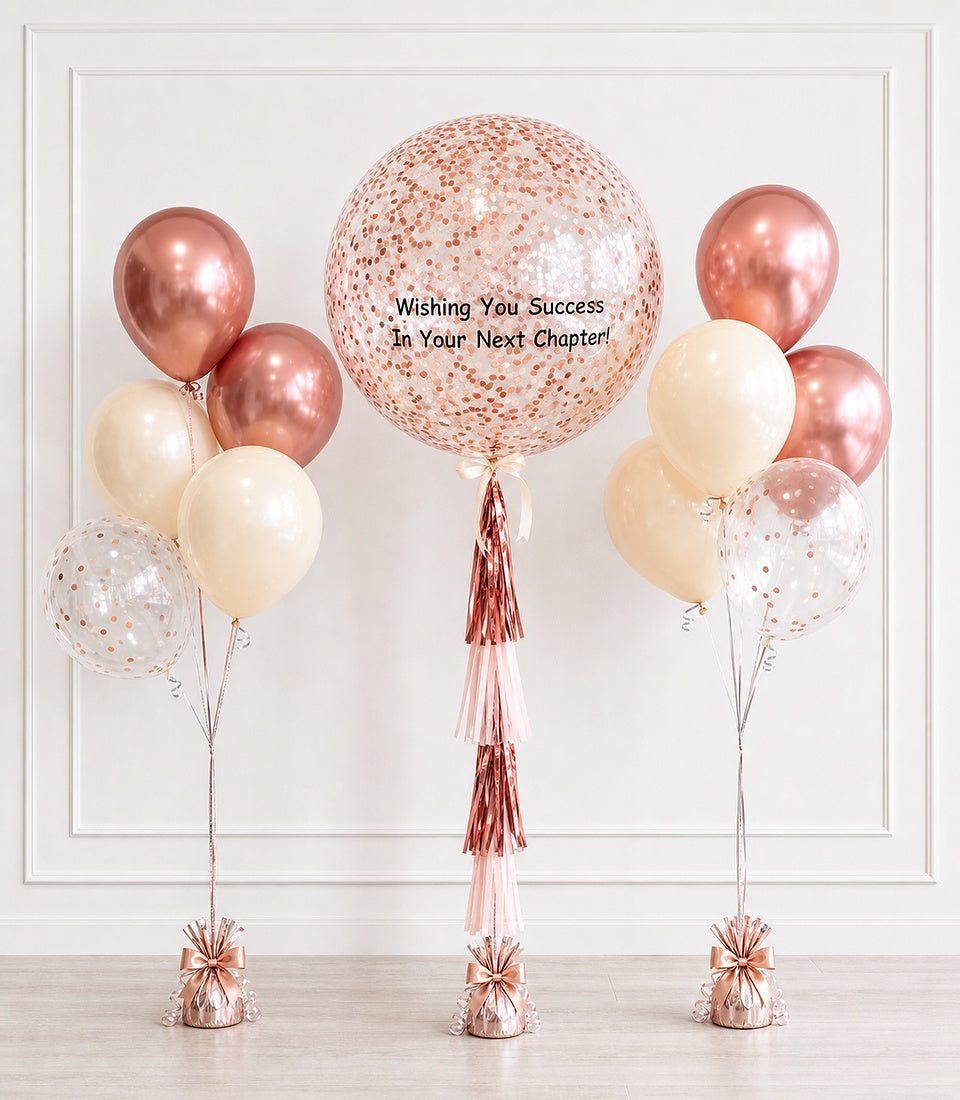 Rose gold and cream balloons with confetti-filled centerpiece and tassels, displaying "Wishing You Success In Your Next Chapter" message.