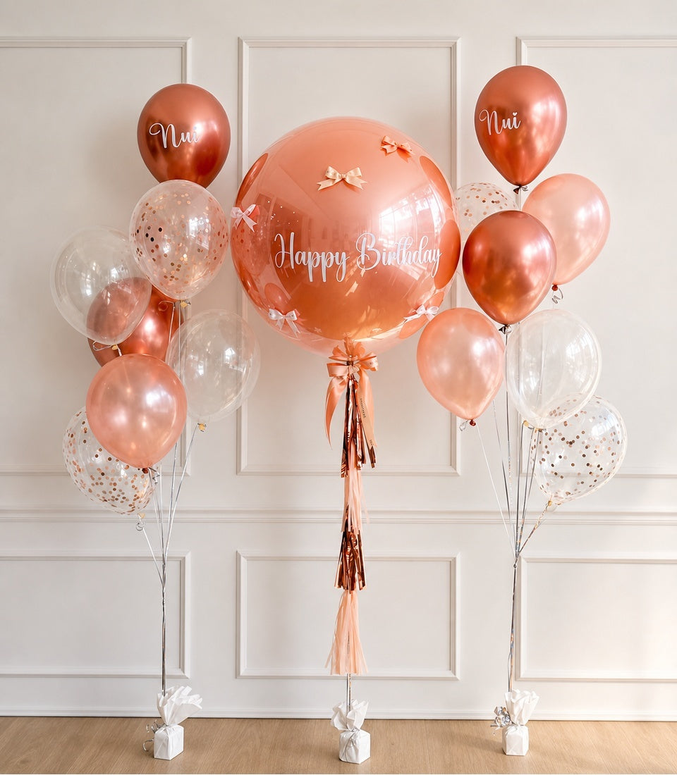 Rose gold and blush balloon bouquet with "Happy Birthday" centerpiece, confetti balloons, personalized names, and tassel decorations against white paneled wall.