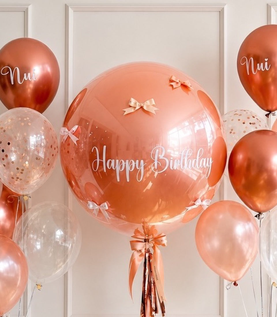 Rose gold birthday balloon arrangement featuring oversized clear bubble balloon with "Happy Birthday" text, metallic balloons, confetti-filled balloons, and ribbon bows.