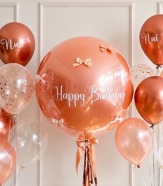 Rose gold birthday balloon arrangement featuring oversized clear bubble balloon with "Happy Birthday" text, metallic balloons, confetti-filled balloons, and ribbon bows.