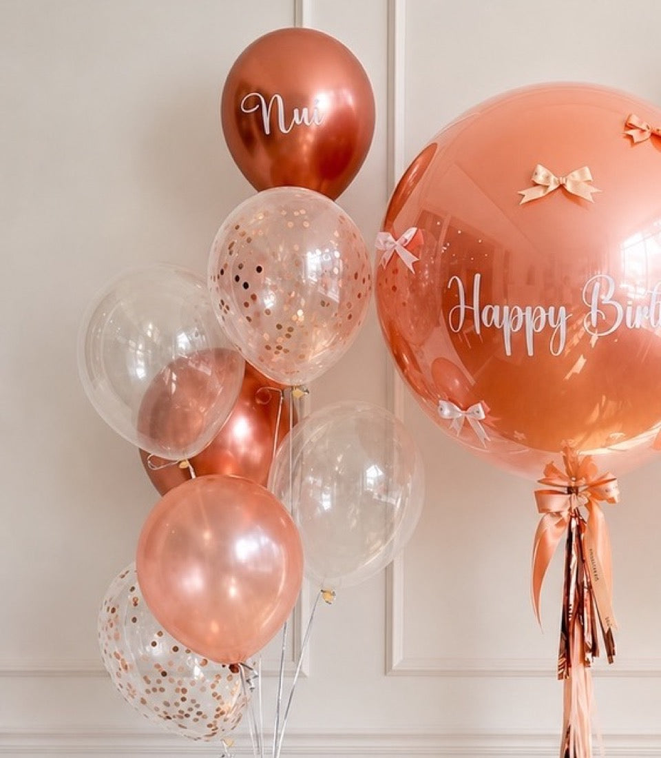 Rose gold and clear balloons with confetti, bows, and "Happy Birthday" text on white wall backdrop.
